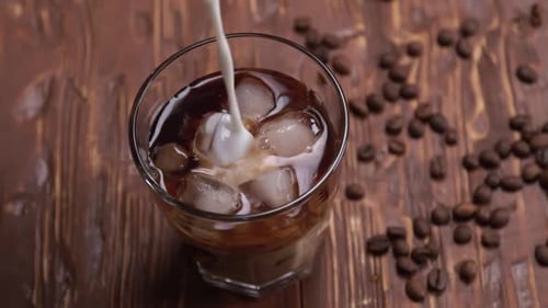 Top View Slow Motion of Cream Being Poured Into a Glass of Cold Brew Iced Coffee on Brown Wood Table