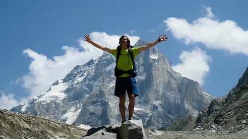 Man Tourist with a Backpack Raises His Hands Up in the Mountains in the Campaign After Climbing