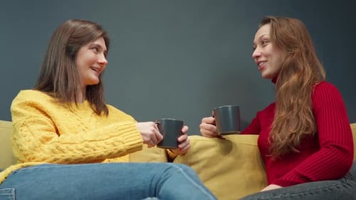 Zwei lächelnde Mädchen diskutieren über Klatsch auf dem gemütlichen gelben Sofa beim Kaffeetrinken
