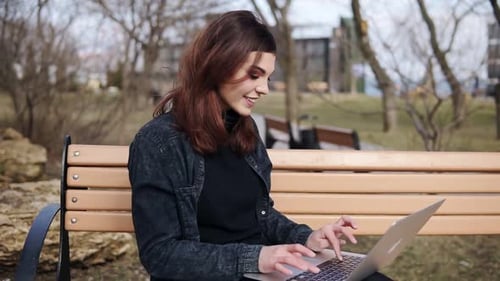 Attractive Female in Her 20'Sis Sitting on the Bench at Park Typing Something on Her Laptop Keyboard