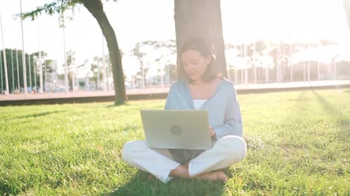 Happy Brunette Woman Using Laptop Computer While Sitting Near the Tree on Grass in Park