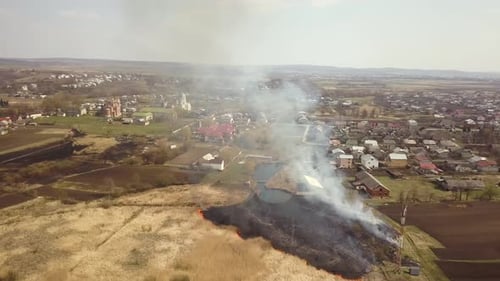 Aerial view of a field with dry grass set on fire with orange flames and high column of smoke.