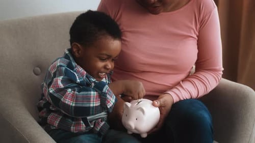Kleiner schwarzer Junge steckt Münzen ins Sparschwein, während er mit Mama sitzt