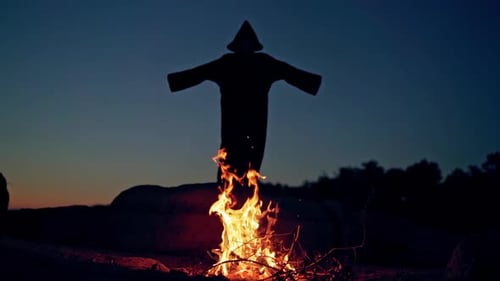 Black death behind the fire flame outdoors. Scary witch in black costume waving arms making fear on