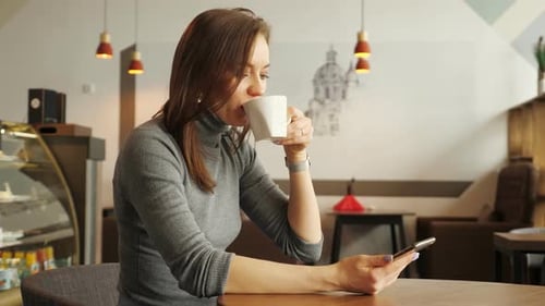 Young Girl in Cafe is Typing a Message on Phone and Drinking a Cup of Coffee