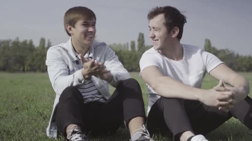 Zwei junge positive Männer sitzen lachend auf einer grünen Sommerwiese