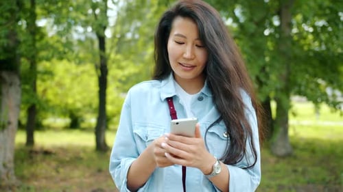 Joyful Asian Girl Using Smartphone Outdoors in City Park Smiling Touching Screen