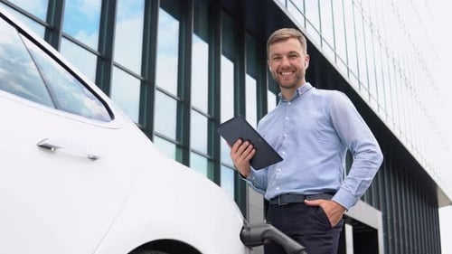 Man Holding Tablet Charging Electric Car Near Office