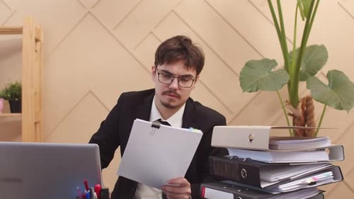 Office Worker Man with Glasses with Serious Look is Working at Desk Looking at Documents and Typing