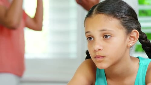 Niña triste sentada a la mesa mientras los padres discuten de fondo
