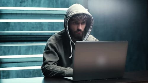 Portrait of Bearded Hacker Man Wearing Sweatshirt with Hood Breaking Password Using Laptop Computer