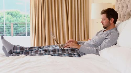 Man Relaxes on Bed Typing on Laptop