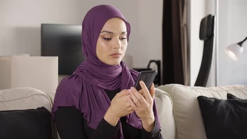 A Young Beautiful Muslim Woman Works on a Smartphone and Smiles at the Camera As She Sits on a Sofa