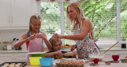 Happy caucasian mother in kitchen with daughter and son, wearing aprons baking cookies together