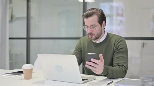 Young Businessman Using Smartphone While Using Laptop in Office