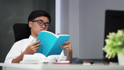 Young Adult Reading Book at Desk in Office