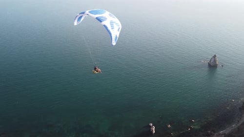 Drohnenaufnahme eines Mannes, der mit einem weißen und blauen Gleitschirm über einen Hügel und Bäume zu den Meereswellen fliegt