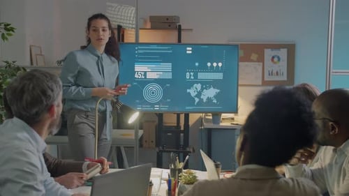 Young Businesswoman Making Presentation for Diverse Group of Colleagues in Dark Office Pointing at