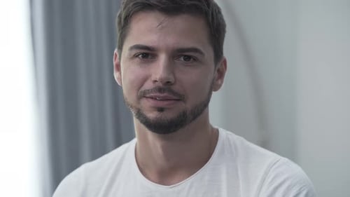 Porträt eines lächelnden Mannes im weißen T-Shirt, der zu Hause sitzt und lächelt, während er in die Kamera schaut