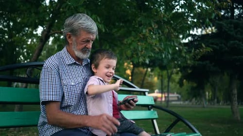 Senior Grandfather and Grandson Are Sitting on a Bench in the Park and Playing on a Smartphone