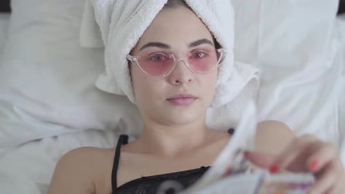 Cute Young Woman in Pink Glasses and Towel on the Head Lying in Bed Reading the Magazine, Journal