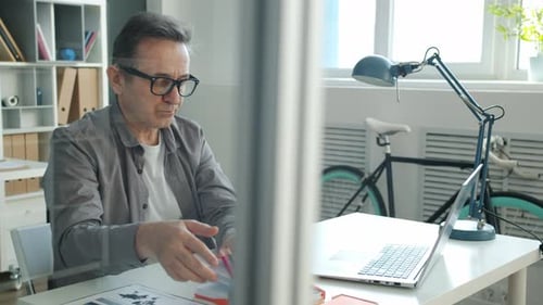 Mature Employee Working in Office Using Laptop Taking Notes in Notebook Alone