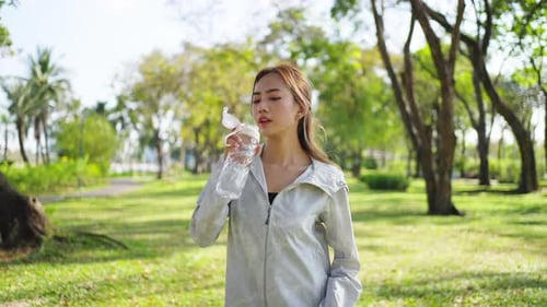 4K Asian woman drinking water from a bottle while jogging at public park in the morning.