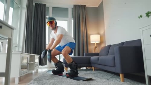 Vídeo divertido. Homem de bermuda e camiseta retrata snowboard em um tapete em um quarto aconchegante. Esperando por