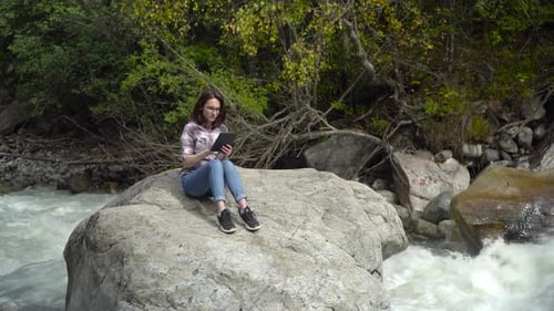 Una mujer joven con gafas se sienta en una piedra con una tablilla en las manos en las montañas cerca del