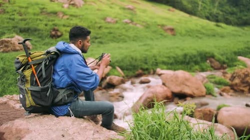 A young male trekker with backpack sits on the rocks of a stream in a green valley.