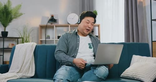 Chico asiático con auriculares disfrutando de su videollamada mientras está sentado en un cómodo sofá