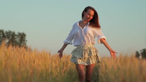 Young Woman in a Skirt and a White Shirt Walks Through a Wheat Field with Hands Outstretched