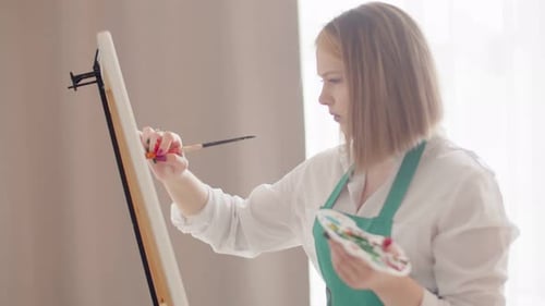Woman is Intently Drawing with Brush on Canvas Holding Palette in Hand Side View