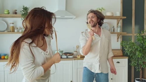 Glückliches Paar tanzt in der Küche und singt Karaoke mit Küchengeräten wie Mikrofonen beim Kochen