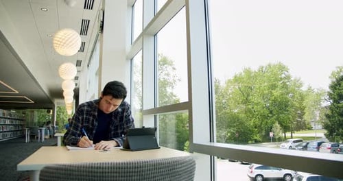 College Student Sitting in Library Writes in a Notebook