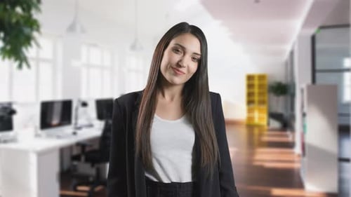 Smiling Young Woman Is Looking at Camera. She Is Standing in a Modern Office in the Background.