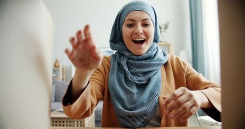 Surprised Middle Eastern Woman in Hijab Opening Carton Box Taking Gift Smiling