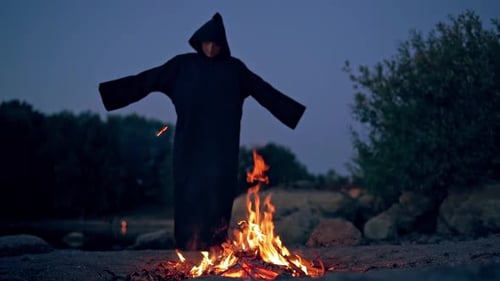 Spooky evening among nature. Dark nightmare by the the fire flame. Black wizard in mantle waving arm