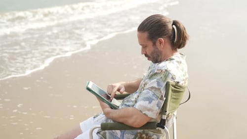 4K Caucasian businessman sitting on outdoor chair and working on digital tablet at the beach.