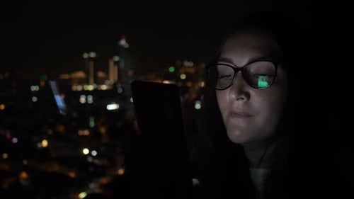 Sleepy Woman Is Flipping Pages on Phone at Night Near the Window with City View.
