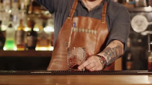 Barman Is Pouring Mixed Drink in a Glass with Big Piece of Ice in a Bar