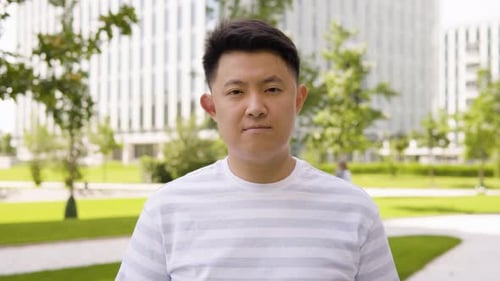 A Young Asian Man Looks Seriously at the Camera in an Urban Area a Park and Office Buildings