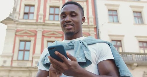 Man in Earrings which Standing Near Urban Ancient Buildings and Texting on Smartphone