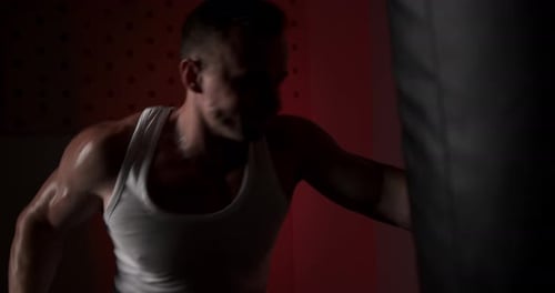 A man in a semi-dark hall, wearing hand wraps, hits a punching bag with quick and accurate punches.