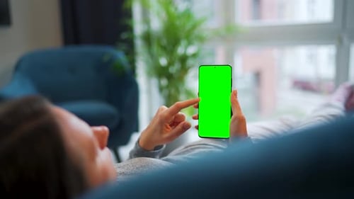 Woman Lies at Home on the Sofa and Using Smartphone with Green Mockup Screen in Vertical Mode