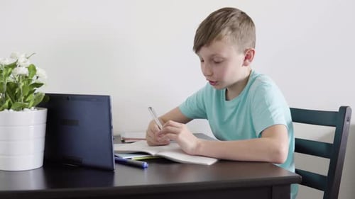 A Young Boy Talks To Someone on a Tablet As He Does Homework for School at a Table at Home