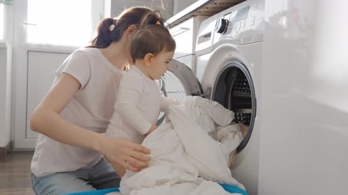 Junge Mutter nimmt Kleidung aus der Waschmaschine mit Baby Girl Kleinkind in der Küche