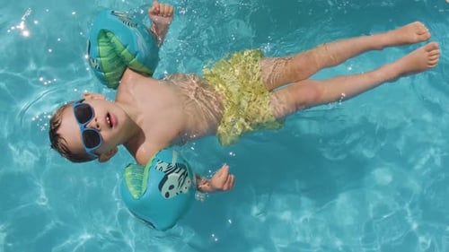 Menino pequeno relaxado usando óculos de sol flutuando na piscina usando flutuadores coloridos