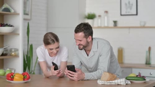 Happy Young Couple Use Phones Together in Kitchen