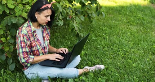 Woman Freelancer in Casual Clothes with Her Computer Laptop Sitting on the Grass at the Park